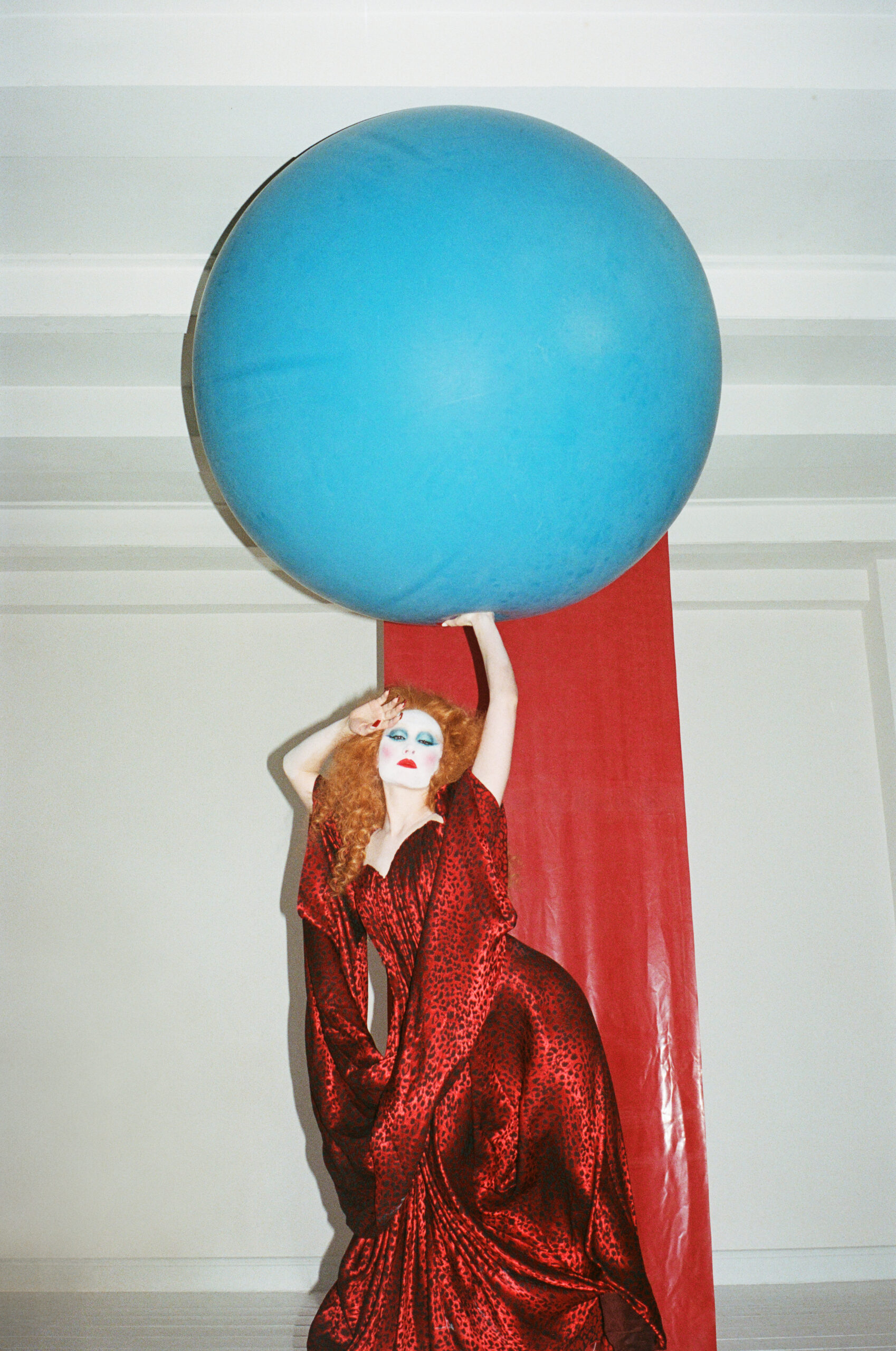 Person in red gown and dramatic makeup poses with one hand on a large blue sphere against a red backdrop.