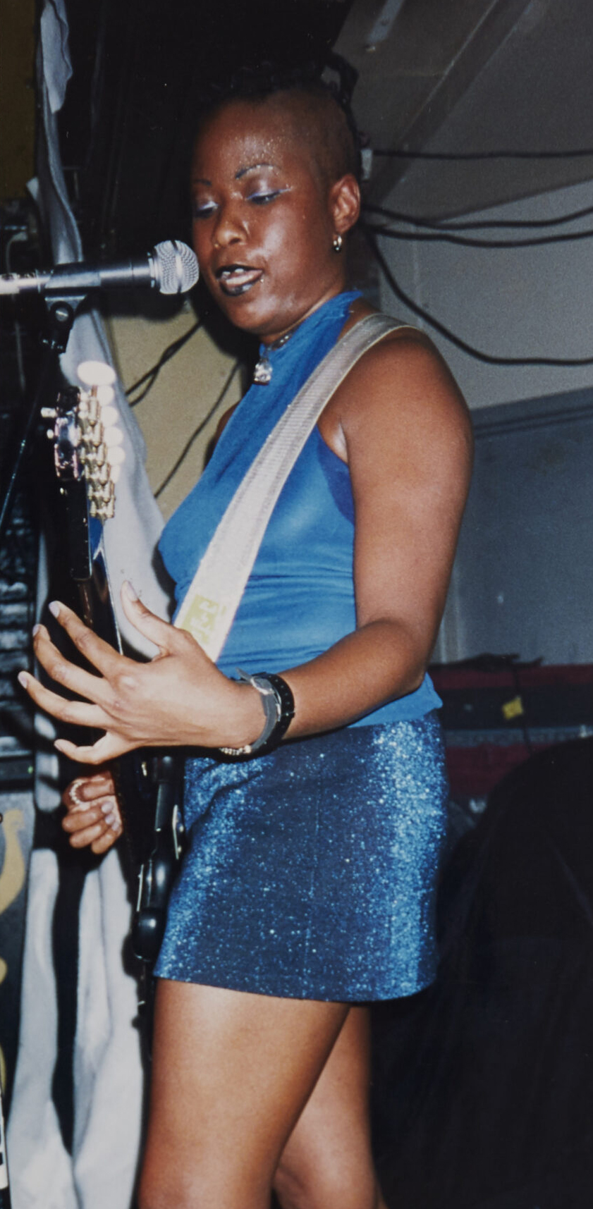 On stage, a performer honors 'How Sista Grrrl Riot Made Room for Black Queers in Punk', singing and playing electric guitar in a blue outfit.